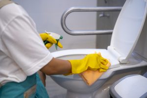 A woman is wiping the toilet with detergent, which is one of the cleaning checklist for schools.