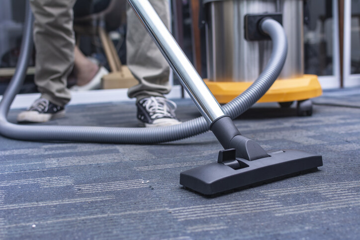 A professional using a heavy-duty industrial vacuum to perform commercial building cleaning on blue patterned office carpeting.