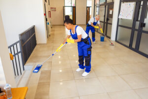 Two professional cleaners in blue overalls mopping a bright hallway, illustrating a key task on a commercial cleaning checklist.