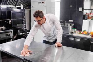 Restaurant cleaning in progress as a commercial kitchen employee sanitizes stainless steel work surfaces to maintain food safety and hygiene standards.